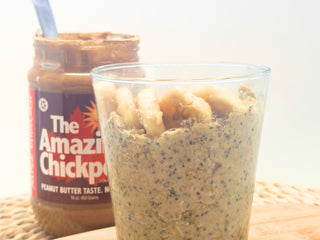 Glass of overnight oats and a jar labeled 'The Amazing Chickpea' on a cutting board against a white background